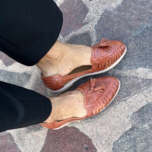 May include: A pair of brown leather sandals with woven straps and tassel accents. The sandals have a comfortable, open-toe design.