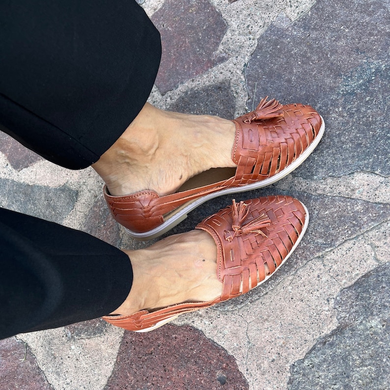 May include: A pair of brown leather sandals with woven straps and tassel accents. The sandals have a comfortable, open-toe design.