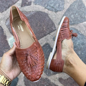 May include: A pair of brown leather sandals with a woven design and a tassel detail. The sandals have a white sole and are open-toe.