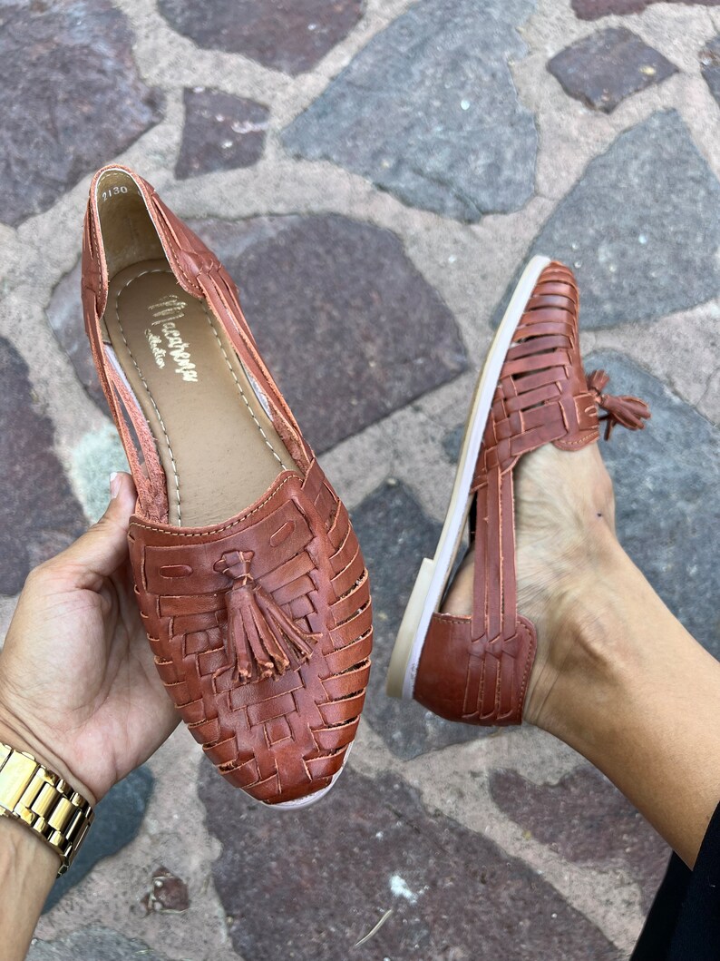 May include: A pair of brown leather sandals with a woven design and a tassel detail. The sandals have a white sole and are open-toe.
