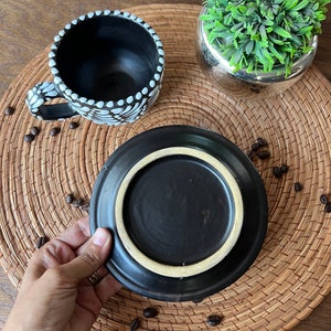 May include: A black ceramic coffee cup with white polka dots and a matching saucer. The cup and saucer are on a woven placemat with coffee beans scattered around them.
