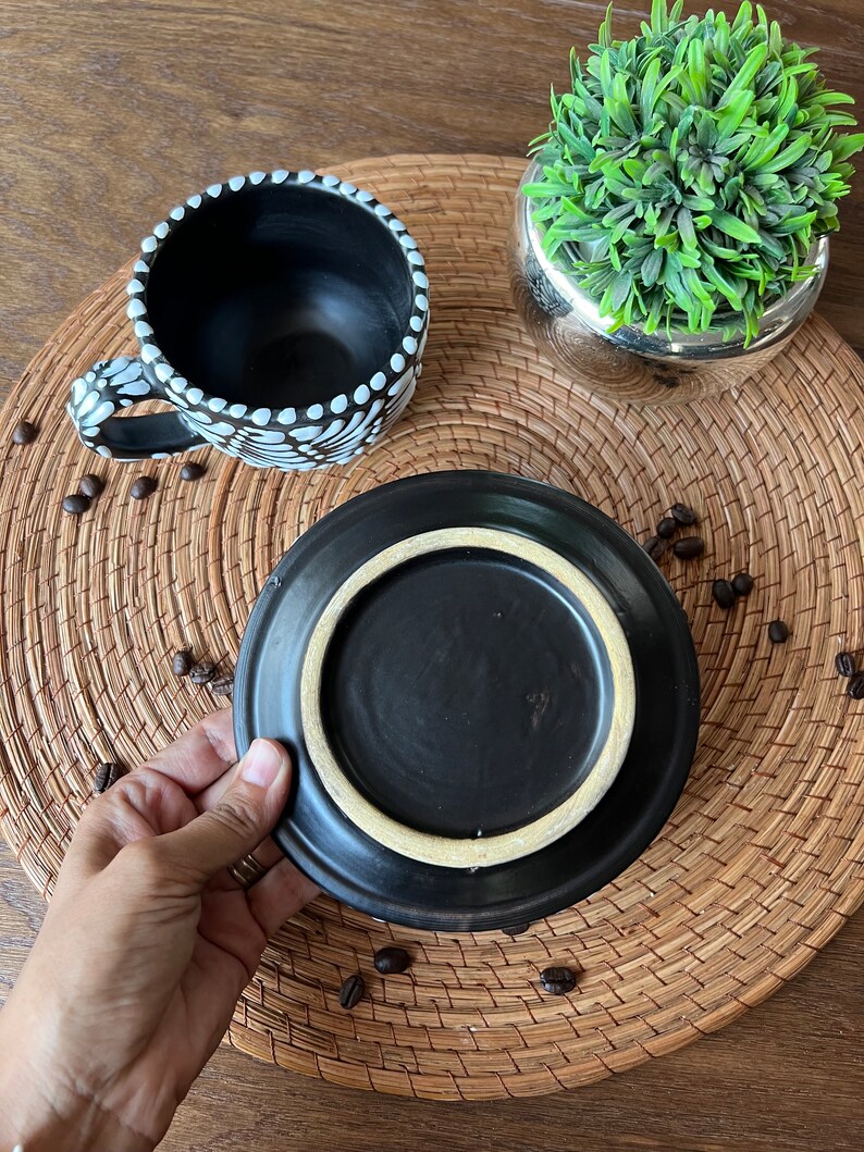 May include: A black ceramic coffee cup with white polka dots and a matching saucer. The cup and saucer are on a woven placemat with coffee beans scattered around them.