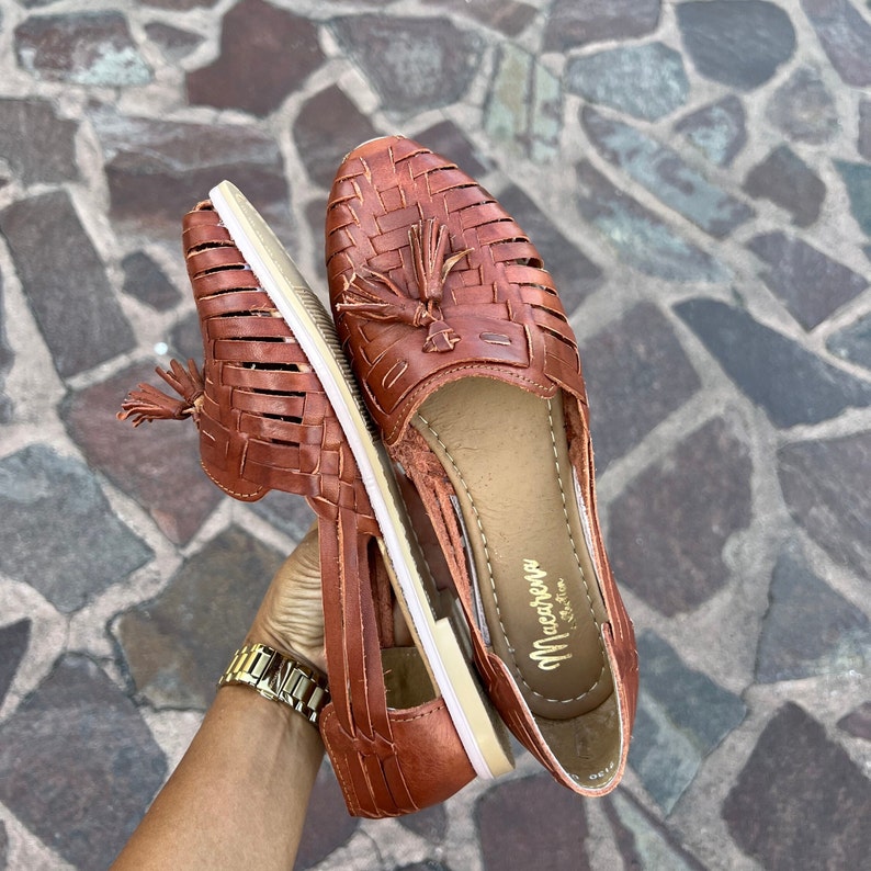 May include: Two pairs of brown leather sandals with woven straps and tassels. The sandals have a white sole and the brand name "Macarena" is printed on the insole.