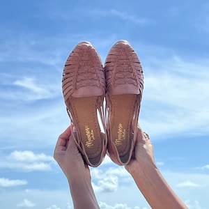 May include: A pair of brown leather sandals with woven straps. The sandals are held up in the air against a blue sky with white clouds.