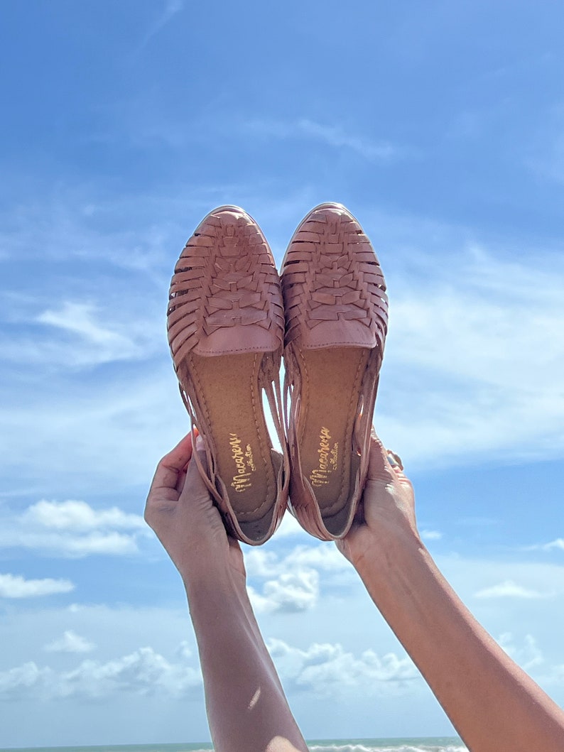 May include: A pair of brown leather sandals with woven straps. The sandals are held up in the air against a blue sky with white clouds.
