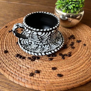 May include: Black and white ceramic coffee cup and saucer with a floral design. The cup and saucer are sitting on a woven placemat with coffee beans scattered around them.