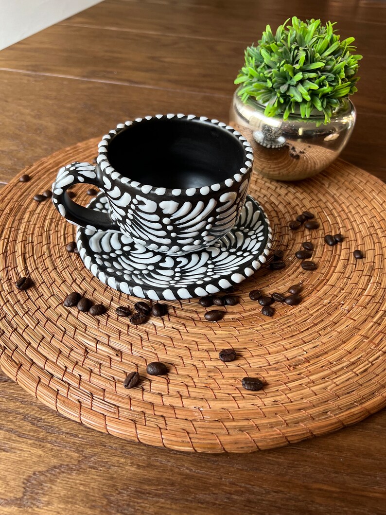 May include: Black and white ceramic coffee cup and saucer with a floral design. The cup and saucer are sitting on a woven placemat with coffee beans scattered around them.
