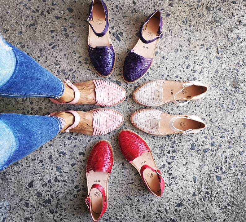 May include: A pair of pink, red, and purple woven leather sandals with ankle straps. The sandals are on a concrete surface.