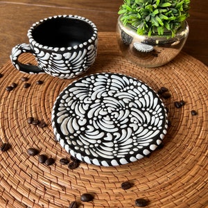 May include: Black and white ceramic coffee cup and saucer with a swirling pattern. The cup and saucer are on a woven placemat with coffee beans scattered around them.