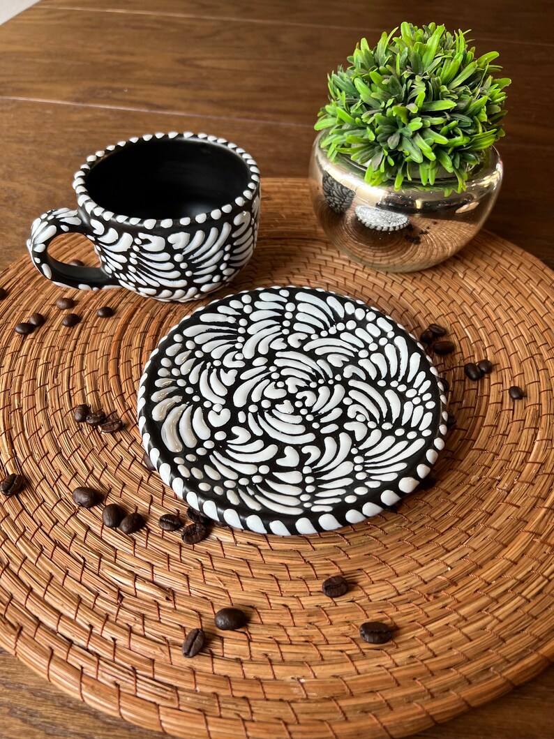 May include: Black and white ceramic coffee cup and saucer with a swirling pattern. The cup and saucer are on a woven placemat with coffee beans scattered around them.