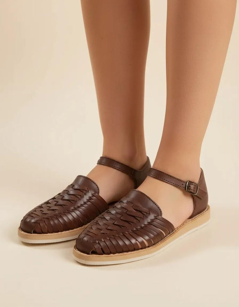 May include: Brown leather sandals featuring a woven design. The sandals have an ankle strap with a buckle and a light-coloured sole. These sandals are designed for comfort and style, suitable for everyday wear.