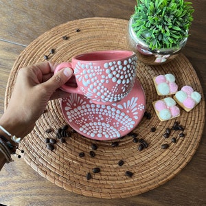 May include: A pink ceramic coffee cup and saucer with a white polka dot design. The cup and saucer are on a woven placemat with coffee beans scattered around them.