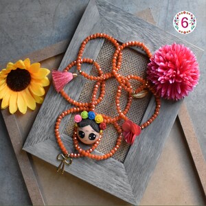 Puede incluir: Un collar de cuentas naranja con un colgante con una cara de dibujos animados con una corona floral. El collar est&aacute; adornado con una borla rosa, un lazo dorado y una flor roja. Un pomp&oacute;n rosa y un girasol amarillo tambi&eacute;n est&aacute;n en la imagen.