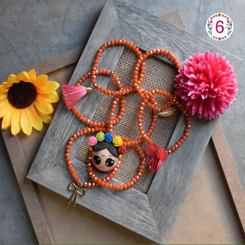 Puede incluir: Un collar de cuentas naranja con un colgante con una cara de dibujos animados con una corona floral. El collar est&aacute; adornado con una borla rosa, un lazo dorado y una flor roja. Un pomp&oacute;n rosa y un girasol amarillo tambi&eacute;n est&aacute;n en la imagen.