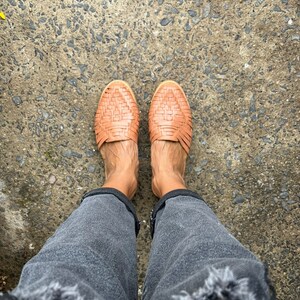 May include: A pair of brown woven leather sandals with a criss-cross pattern. The sandals are worn on a person's feet, with the person wearing dark gray jeans.