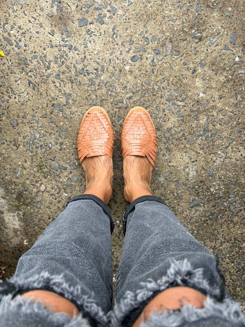 May include: A pair of brown woven leather sandals with a criss-cross pattern. The sandals are worn on a person's feet, with the person wearing dark gray jeans.