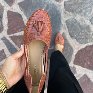 May include: Brown leather sandals with a woven design and a tassel. The sandals have a cut-out design on the sides and the brand name "Paca&ntilde;a" is visible on the insole.