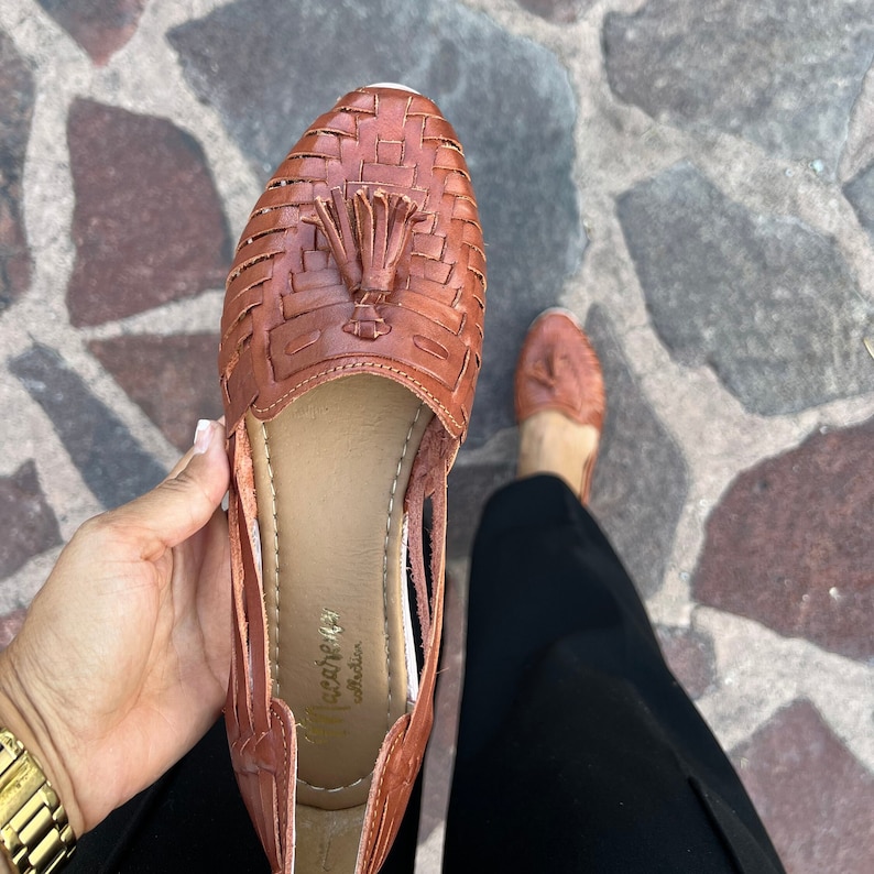 May include: Brown leather sandals with a woven design and a tassel. The sandals have a cut-out design on the sides and the brand name "Paca&ntilde;a" is visible on the insole.