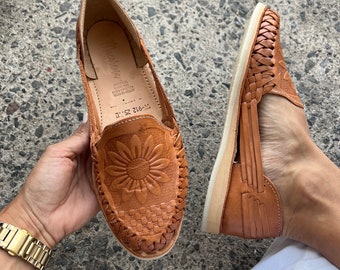 May include: A pair of brown leather sandals with a woven design. The sandals have a sunflower design on the front and a woven strap across the top.