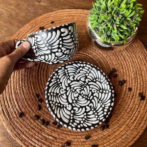 May include: A black and white ceramic coffee cup and saucer set with a swirling floral pattern. The set is on a woven placemat with coffee beans scattered around it.
