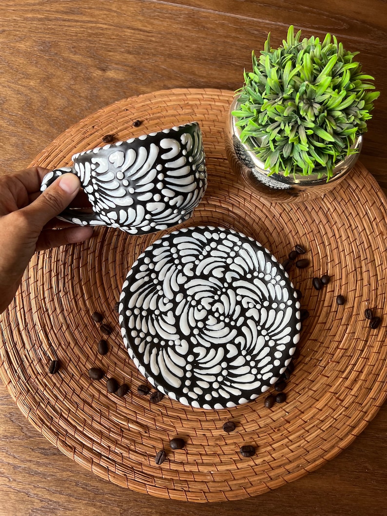 May include: A black and white ceramic coffee cup and saucer set with a swirling floral pattern. The set is on a woven placemat with coffee beans scattered around it.