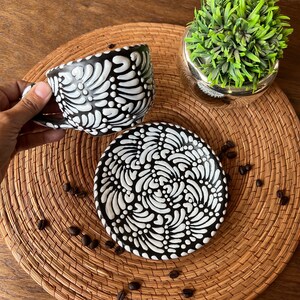 May include: Black and white ceramic coffee cup and saucer set with a swirling floral design. The cup and saucer are on a woven placemat with coffee beans scattered around them.