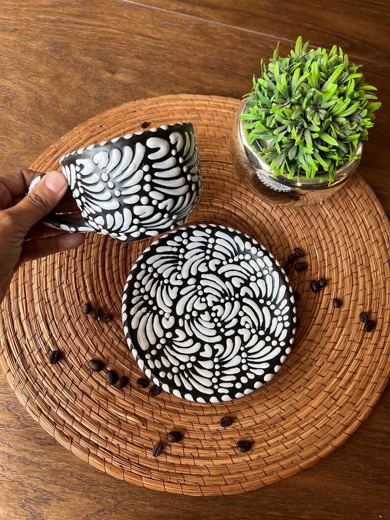 May include: Black and white ceramic coffee cup and saucer set with a swirling floral design. The cup and saucer are on a woven placemat with coffee beans scattered around them.