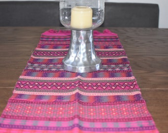 Op de afbeelding: Een roze en grijze tafelrunner met een geometrisch patroon. Een zilveren kaarsenhouder met een transparante glazen bovenkant staat op de runner.