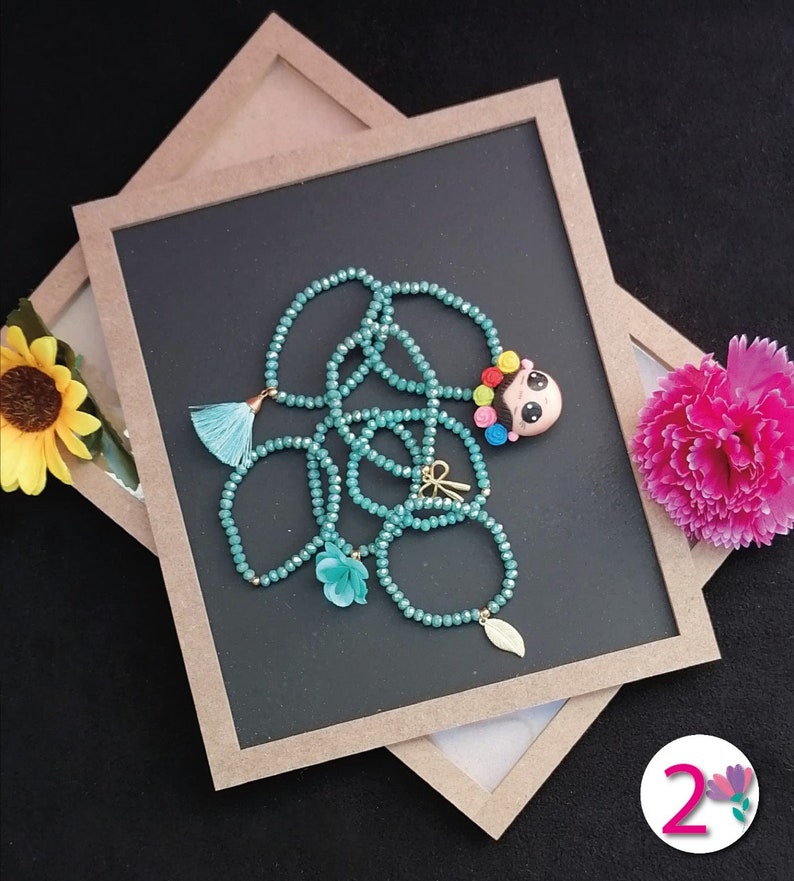 Puede incluir: Un collar de cuentas turquesas con borla, flor y dije de hoja, junto con un colgante de una ni&ntilde;a con cabello colorido. El collar se exhibe sobre una superficie negra con un marco marr&oacute;n y flores decorativas.