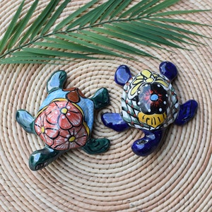 May include: Two hand-painted ceramic turtles with colourful floral designs. The turtle on the left is green with orange, pink, and yellow flowers. The turtle on the right is blue with yellow, black, and red flowers.