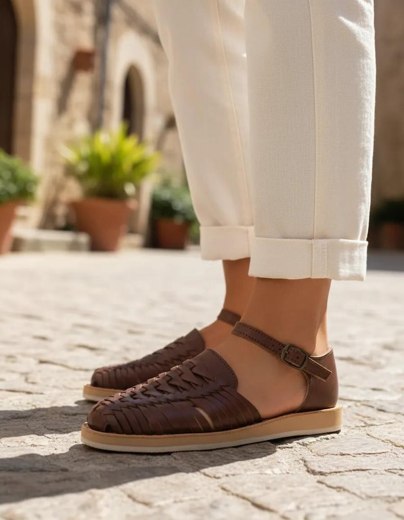 May include: Brown leather sandals with woven detailing and a buckle fastening. The sandals have a flat sole and are worn with cream trousers. The image is taken outdoors on a cobbled surface.