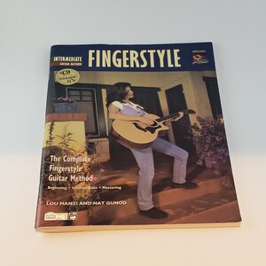 May include: A book titled "Fingerstyle: The Complete Fingerstyle Guitar Method" by Lou Manzi and Nat Gunod. The book cover features a woman playing an acoustic guitar on a porch.