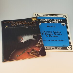 May include: Two guitar instruction books. One book is titled "Intros, Endings & Turnarounds" with a guitar image. The other book is titled "Didactic Series for Guitar Book 2" with blue cover. Both books are on a white surface.