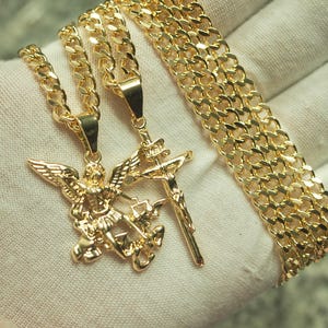 May include: Gold-colored necklaces and a bracelet are displayed. One necklace features an angel pendant, the other a crucifix. The bracelet and necklaces have a curb chain design. The jewelry is presented on a white surface.