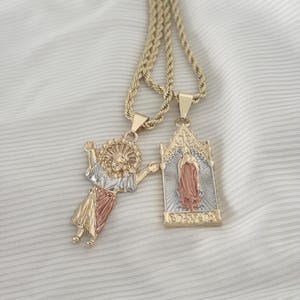 Puede incluir: Dos collares dorados con colgantes religiosos. Uno representa una figura con los brazos extendidos, y el otro presenta un icono religioso dentro de un marco rectangular. Las cadenas tienen un diseño de cuerda.