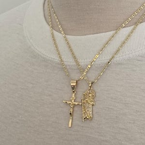May include: Two gold-colored necklaces with pendants. One pendant is a crucifix, and the other depicts a religious figure. The necklaces are displayed on a white t-shirt.