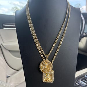May include: A gold-toned double-layered chain necklace featuring two pendants. The top pendant is circular with a raised figure, and the bottom pendant is rectangular with a central figure and radiating lines, both suspended from a curb chain.