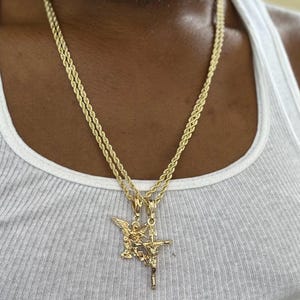 May include: A gold rope chain necklace with a pendant featuring an angel holding a cross. The pendant is made of two-toned gold.