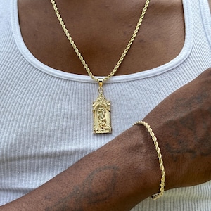 May include: A gold rope chain necklace with a pendant featuring a religious figure. The necklace is worn by a person with dark skin. The person is also wearing a gold rope chain bracelet on their wrist.