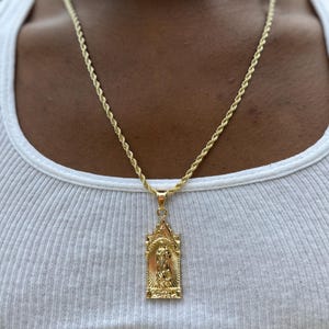 Op de afbeelding: Een gouden touwketting met een hanger met een religieus figuur in een gouden frame.