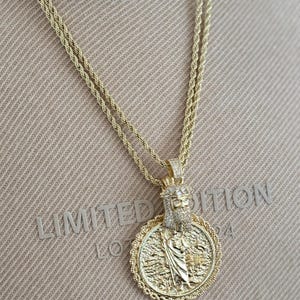 Pode incluir: Colar com pingente dourado com um design detalhado de figura religiosa. O pingente é adornado com detalhes brilhantes e pende de uma corrente dupla de corda. O fundo exibe o texto "LIMITED EDITION".