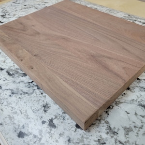 May include: A rectangular wooden cutting board with a natural wood grain finish. The board is made of a dark brown wood and has a smooth, polished surface. The edges are cleanly cut, and the board appears to be new.