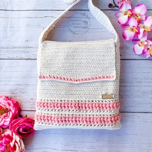 May include: A white crocheted crossbody bag with a pink trim and a pink and white crocheted design. The bag has a strap and a small label that says "Chunky Hunky Creations".