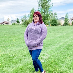 May include: A woman wearing a purple crocheted hooded sweater with a pocket in the front, blue jeans, and sandals. She is standing in a grassy field.