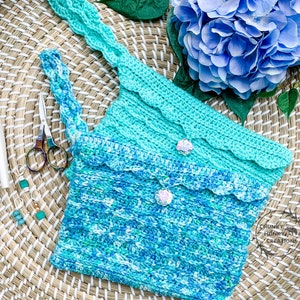 May include: A crocheted bag with a blue and green ombre design. The bag has a scalloped edge and a small, white button closure. The bag is sitting on a woven basket with two crochet hooks and a pair of scissors.