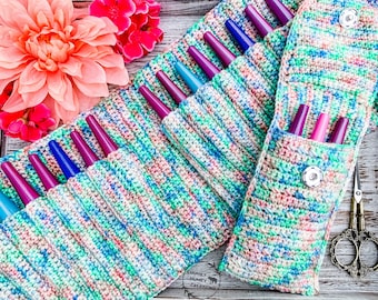 May include: A crocheted multi-coloured case with multiple pockets for holding knitting needles. The case is made with a variegated yarn in shades of blue, green, pink, and white. The case has a button closure and is lying on a wooden surface.