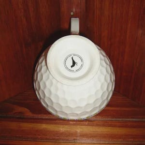 Peut inclure: Tasse en c&eacute;ramique blanche avec une texture de balle de golf. La tasse a une anse et un logo circulaire en bas. La tasse est pos&eacute;e sur un fond en bois.