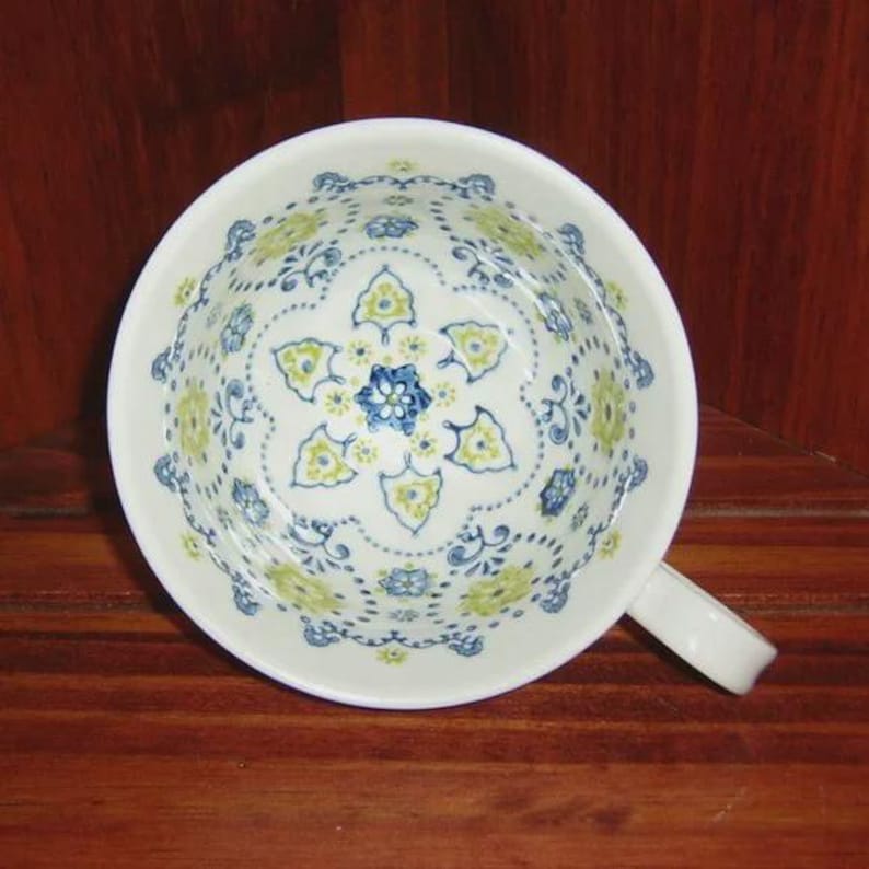 Peut inclure: Tasse &agrave; th&eacute; en c&eacute;ramique blanche avec une anse, orn&eacute;e d'un motif floral bleu et jaune. Le motif comprend une fleur centrale et des &eacute;l&eacute;ments d&eacute;coratifs sur le bord. La tasse est pos&eacute;e sur une surface en bois.