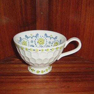 Peut inclure: Tasse &agrave; th&eacute; blanche avec une anse et un motif floral d&eacute;coratif bleu et vert. La tasse a un ext&eacute;rieur textur&eacute; et nervur&eacute; et repose sur un petit socle. La tasse &agrave; th&eacute; est pr&eacute;sent&eacute;e sur une surface en bois.
