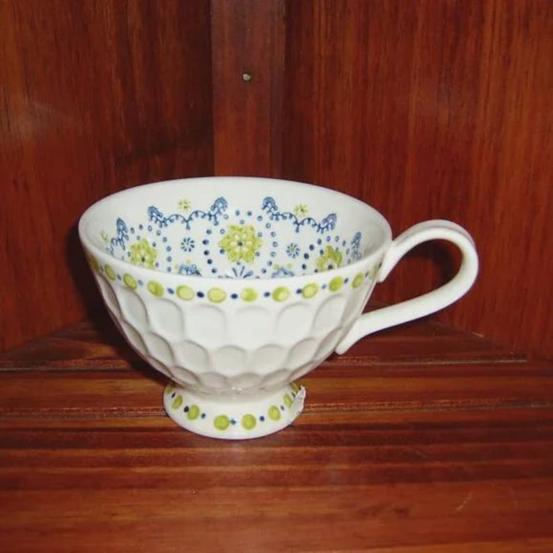 Peut inclure: Tasse &agrave; th&eacute; blanche avec une anse et un motif floral d&eacute;coratif bleu et vert. La tasse a un ext&eacute;rieur textur&eacute; et nervur&eacute; et repose sur un petit socle. La tasse &agrave; th&eacute; est pr&eacute;sent&eacute;e sur une surface en bois.
